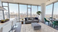 This high-rise residence is defined by its dramatic floor-to-ceiling glass walls, which wrap around the living and dining areas to deliver unobstructed panoramic views of a sprawling city skyline. Dark hardwood flooring runs throughout the open-plan living space, grounding the bright, airy interior and providing a warm contrast to the white walls and abundant natural light that pours in throughout the day.

The living area is generously proportioned, accommodating a full seating arrangement alongside a dedicated dining space with a round table set against the expansive glass perimeter. A structural cylindrical column adds an architectural accent to the corner of this combined space. The kitchen runs in a clean galley configuration, outfitted with white flat-front cabinetry, quartz countertops, a stainless steel undermount sink, and stainless steel appliances, with a floor-to-ceiling window at the end framing additional skyline views.

The bedrooms are calm and well-lit, each benefiting from large windows overlooking the cityscape. One bedroom includes a dedicated closet and direct access to a full bathroom. The bathrooms are finished with large-format marble-look tile, wood-toned vanities, white quartz countertops, backlit mirrors, and glass-enclosed walk-in showers, conveying a clean, contemporary aesthetic throughout.
