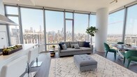 This high-rise residence is defined by its dramatic floor-to-ceiling glass walls, which wrap around the living and dining areas to deliver unobstructed panoramic views of a sprawling city skyline. Dark hardwood flooring runs throughout the open-plan living space, grounding the bright, airy interior and providing a warm contrast to the white walls and abundant natural light that pours in throughout the day.

The living area is generously proportioned, accommodating a full seating arrangement alongside a dedicated dining space with a round table set against the expansive glass perimeter. A structural cylindrical column adds an architectural accent to the corner of this combined space. The kitchen runs in a clean galley configuration, outfitted with white flat-front cabinetry, quartz countertops, a stainless steel undermount sink, and stainless steel appliances, with a floor-to-ceiling window at the end framing additional skyline views.

The bedrooms are calm and well-lit, each benefiting from large windows overlooking the cityscape. One bedroom includes a dedicated closet and direct access to a full bathroom. The bathrooms are finished with large-format marble-look tile, wood-toned vanities, white quartz countertops, backlit mirrors, and glass-enclosed walk-in showers, conveying a clean, contemporary aesthetic throughout.