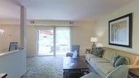 This single-floor residence offers a welcoming and well-appointed living space designed for comfort and ease of living. The living room serves as the heart of the home, featuring a spacious, open layout anchored by a plush sofa and complementary seating. A large, dark wood coffee table grounds the seating arrangement over a subtly patterned area rug, adding warmth and definition to the space.

One of the most notable features of the living area is the expansive sliding glass door that spans a significant portion of the exterior wall. This architectural element floods the room with natural light, creating a bright and airy atmosphere throughout the day. The doors provide a seamless transition to an outdoor patio area, where lush greenery and outdoor furnishings are visible, suggesting an inviting extension of the living space.

The interior walls are finished in a soft, neutral palette that enhances the sense of openness and light. Ceiling-mounted HVAC vents indicate a central climate control system for year-round comfort. Accent lighting, including a classic table lamp, adds a warm, layered glow to the room during evening hours. Overall, the space combines functional design with a calm, understated aesthetic.
