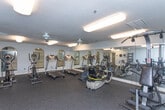 Located at 2761 Woodland Terr SE in Smyrna, Georgia, this single-floor fitness center is designed to provide a well-rounded workout environment for its users. The space is thoughtfully organized into distinct functional zones, all unified by durable rubber flooring and soft blue-gray walls that contribute to a clean, focused atmosphere.

The primary exercise area is generously sized and features a row of treadmills aligned along a full-length mirrored wall with arched accent mirrors, creating a sense of expanded space and allowing users to monitor their form. Ceiling-mounted fluorescent fixtures provide bright, even lighting throughout. Ceiling fans help maintain comfortable airflow during workouts.

Complementing the cardio section, the room also accommodates a multi-station cable machine for strength training, alongside elliptical trainers and a recumbent cycle. A freestanding ladder-style shelf holds free weights and accessories, paired with a cushioned bench for seated exercises. Wall-mounted dispensers suggest additional amenity provisions for users.

A secondary entry or transitional area features double panel doors, additional rubber flooring, and a small bench, serving as a practical buffer zone between the fitness space and the exterior. Textured ceilings throughout add architectural consistency to the space. Overall, the fitness center offers a comprehensive and functional environment suitable for a wide range of exercise routines.