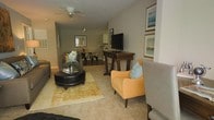 This single-floor space offers an open-concept layout that seamlessly connects the living and dining areas, creating a cohesive and functional flow throughout. The living area features plush carpeting underfoot and is furnished with a mix of seating options, including a warm amber accent chair with a decorative blue pillow and a tufted leather ottoman atop a golden area rug, adding layers of texture and color to the space. A wooden console table serves as an entertainment stand, housing a flat-screen television and providing additional surface space. The walls are painted in soft, neutral tones and adorned with framed artwork featuring calming, nature-inspired imagery, contributing to the space's relaxed ambiance. A ceiling fan overhead provides both practical ventilation and a subtle design element. The dining area, visible in the background, accommodates a table and chairs beneath a track lighting fixture, while a glimpse of cabinetry hints at a kitchen beyond. Natural light filters in through a window in the dining area, brightening the space. Overall, the layout and furnishings create a comfortable, well-appointed living environment with a cohesive, contemporary aesthetic.