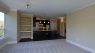 This single-floor space showcases an open-concept layout that integrates the kitchen and living area into one cohesive environment. The kitchen serves as a focal point, featuring dark espresso cabinetry that contrasts elegantly with stainless steel appliances, including a refrigerator, microwave, and range. A substantial dark kitchen island anchors the space, while three pendant lights hang above it, providing targeted task lighting and a stylish visual element. Recessed lighting throughout the kitchen adds further illumination.

Crown molding frames the ceilings, lending a refined architectural detail to the interior. The flooring transitions from dark hardwood in the kitchen zone to plush neutral carpet in the living area, clearly delineating each functional space. Built-in shelving units along the living area wall offer practical display and storage opportunities.

A hallway visible in the background suggests additional rooms beyond, with a wall sconce providing warm accent lighting. The overall ambiance is contemporary and polished, with a carefully balanced palette of warm neutrals, dark tones, and metallic accents creating a sophisticated, welcoming atmosphere.