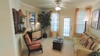 This single-floor space offers a warm and inviting atmosphere, highlighted by soft neutral wall tones and plush carpeting throughout. The living area serves as the focal point of the space, featuring crown molding that adds a refined architectural touch along the ceiling perimeter. A ceiling fan with an integrated light fixture provides both comfort and ambient lighting, complementing the natural light that filters in through large windows dressed with sheer orange curtains, lending a warm, golden quality to the room.

A full-light exterior door offers direct access to the outdoors while allowing additional natural light to brighten the interior. The living area comfortably accommodates seating arrangements, with space for accent chairs and a central coffee table, creating a functional and relaxed gathering area. A decorative dresser and framed wall art contribute to the curated, homey aesthetic of the room.

Lush indoor plants add a natural, organic element to the space, enhancing its welcoming ambiance. The overall design balances practicality with comfort, making it a well-appointed space suited for everyday living.