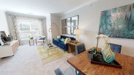 This single-floor residence is thoughtfully designed with a cohesive color palette of navy blue, gold, and teal accents that carry throughout the space, creating a polished and contemporary ambiance. Crown molding detail adds architectural refinement to the main living area, which features soft carpeting, large windows with decorative curtains, and ample natural light. The living room is anchored by a deep blue velvet sofa, a glass-top coffee table, and a gallery-style arrangement of framed mirrors that visually expand the room. A dedicated dining area sits adjacent, furnished with a rich wood table and upholstered seating. The kitchen is equipped with stainless steel appliances, dark granite countertops, white cabinetry, and wood-style flooring, offering both function and style. The bedroom features a dramatic navy accent wall, plush carpeting, a ceiling fan, and generous natural light through patterned curtains, creating a serene retreat. The bathroom includes dark hardwood-style flooring, an extended white vanity with dark granite counters, an arched alcove housing a soaking tub with tile surround, and a large mirror with contemporary lighting — combining practical storage with refined design.