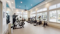 This fitness center is designed to accommodate a full range of workout needs within a single, open-plan space. The room's most distinctive architectural feature is its tray ceiling, painted a bold teal blue and accented with recessed lighting and a ceiling fan, creating a visually dynamic contrast against the crisp white walls. The elevated ceiling height contributes to an open, airy atmosphere throughout.

Large windows line the perimeter walls, allowing generous amounts of natural light to flood the interior while offering views of the surrounding outdoor area. The durable, speckled flooring provides a practical and clean surface suited for heavy equipment and high-traffic use.

The cardio zone is well-appointed with multiple treadmills, elliptical trainers, and stationary and spin bikes arranged in organized rows along the windowed wall. The strength training area features a multi-station cable machine, a Smith machine, free weight racks stocked with a range of dumbbells, adjustable benches, and stability balls. Large wall mirrors are strategically placed to visually expand the space and assist users with form. A wall-mounted flat-screen television is also present. The layout efficiently separates cardio and strength zones, maximizing usability across the entire floor.
