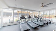 This fitness center is thoughtfully designed to support a comprehensive workout experience. The cardio zone is defined by a floor-to-ceiling glass wall that floods the space with natural light, offering views of an outdoor pergola area and landscaped surroundings. Multiple treadmills and an elliptical machine are arranged along this window wall, creating an open, airy environment for cardio training. Ceiling fans overhead provide additional ventilation and comfort during workouts.

The strength training area occupies the interior portion of the center, featuring soft green accent walls and recessed lighting. A variety of resistance machines are positioned throughout, including cable systems, chest and leg press stations, a rowing machine, and adjustable benches. A wall-mounted flat-screen television adds entertainment options for users during their sessions. Wall-mounted accessory storage keeps the space organized and functional.

A dedicated functional training zone provides additional flexibility, equipped with a multi-functional training tower and resistance band attachments mounted to the wall. The entire space is finished with dark, commercial-grade rubber flooring, ideal for both equipment stability and user safety. The overall layout is clean, organized, and designed to accommodate a wide range of fitness activities in a comfortable community setting.
