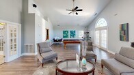 Located in Washington Quarters in Avon, Indiana, this single-floor community clubhouse offers a well-appointed gathering space designed for both relaxation and recreation. The interior is defined by striking vaulted ceilings that lend an open, airy feel throughout. Neutral gray walls and warm hardwood-style flooring create a cohesive and welcoming aesthetic across the entire space.

The main lounge area serves as the heart of the clubhouse, anchored by a tile-surround fireplace topped with a wall-mounted flat-screen television. Comfortable upholstered seating is arranged around a patterned area rug, encouraging conversation and relaxation. A large arched window and French doors flood the room with abundant natural light.

Adjacent to the lounge is a recreation area featuring a full-sized billiards table, additional seating, vibrant wall art, and a ceiling fan — ideal for casual social gatherings. A dining area with sleek seating is also visible in this zone.

The kitchenette is equipped with white cabinetry, a granite-topped breakfast bar with modern bar stools, pendant lighting, tile flooring, and standard appliances, making it convenient for community events and catering needs.

French doors with sidelights connect the spaces seamlessly, enhancing the flow and openness of the clubhouse while allowing natural light to penetrate throughout.