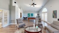 Located in Washington Quarters in Avon, Indiana, this single-floor community clubhouse offers a well-appointed gathering space designed for both relaxation and recreation. The interior is defined by striking vaulted ceilings that lend an open, airy feel throughout. Neutral gray walls and warm hardwood-style flooring create a cohesive and welcoming aesthetic across the entire space.

The main lounge area serves as the heart of the clubhouse, anchored by a tile-surround fireplace topped with a wall-mounted flat-screen television. Comfortable upholstered seating is arranged around a patterned area rug, encouraging conversation and relaxation. A large arched window and French doors flood the room with abundant natural light.

Adjacent to the lounge is a recreation area featuring a full-sized billiards table, additional seating, vibrant wall art, and a ceiling fan — ideal for casual social gatherings. A dining area with sleek seating is also visible in this zone.

The kitchenette is equipped with white cabinetry, a granite-topped breakfast bar with modern bar stools, pendant lighting, tile flooring, and standard appliances, making it convenient for community events and catering needs.

French doors with sidelights connect the spaces seamlessly, enhancing the flow and openness of the clubhouse while allowing natural light to penetrate throughout.