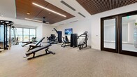 Located on Village Pkwy in Flower Mound, TX, this contemporary fitness center offers a single, thoughtfully designed exercise room that provides ample space for a full range of workout activities. The room is defined by its striking architectural ceiling, which features warm wood slat paneling set within a recessed, coffered framework, complemented by integrated linear lighting and a ceiling fan that contribute to a comfortable and inviting atmosphere.

The floor is finished with neutral, textured carpet tiles that span the entire space, lending a clean and modern aesthetic. Large floor-to-ceiling glass doors and windows along one wall flood the interior with natural light and offer direct views of an outdoor terrace area, where palm trees and open-air seating are visible, seamlessly connecting the interior to the exterior.

The facility is comprehensively equipped with a wide array of strength training machines, a Smith machine, adjustable benches, and a full rack of dumbbells organized along a mirrored wall. The mirrored surfaces enhance the sense of openness and allow users to monitor their form. A wall-mounted flat-screen TV adds a media element for entertainment during workouts. A built-in storage and counter area near the entrance provides functional utility. Overall, the space delivers a polished, high-end fitness environment suited for serious training.