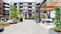 This two-level outdoor courtyard is designed as a comprehensive amenity hub, anchored by a generously sized swimming pool with fountain features and a surrounding deck outfitted with lounge chairs and umbrellas. The pool area is complemented by additional seating clusters, offering residents multiple zones for relaxation and socializing.

The courtyard's architecture blends dark gray siding with warm red and wood-toned accents, creating a contemporary yet inviting aesthetic. A prominent pergola structure with heavy timber beams and steel supports defines a shaded lounge area furnished with upholstered seating, providing a comfortable outdoor retreat. Adjacent to this, a long communal bar with high stools serves as a central gathering point.

A built-in outdoor kitchen, constructed with natural stacked stone, features multiple grills and is nestled alongside raised planters filled with lush greenery and flowering plants. Manicured landscaping throughout softens the hardscape and enhances the sense of a well-maintained, resort-style environment. The courtyard is enclosed by the multi-story building on multiple sides, offering a sense of privacy and seclusion while maintaining open-air exposure to natural light.