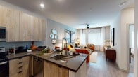 This single-floor residence showcases a thoughtfully designed open-concept layout that seamlessly connects the kitchen, dining, and living areas. The kitchen is a standout feature, outfitted with light wood cabinetry extending to the ceiling, dark quartz countertops, stainless steel appliances including a refrigerator, electric range, microwave, and dishwasher, and a peninsula island with an integrated sink — ideal for casual dining and meal preparation. Wood-look flooring runs throughout the main living areas, adding warmth and continuity to the space. The living room is bright and welcoming, benefiting from natural light through large windows dressed with white curtains and blinds. A ceiling fan overhead enhances comfort, while the soft blue-grey accent wall adds a touch of contemporary character. The bedroom offers a serene retreat with plush carpeting, a large window, and a soft sage-green accent wall. The bathroom features wood-tone vanity cabinetry, dark countertops, a large mirror, and a tub-shower combination with a stylish curtain. Recessed lighting throughout the unit ensures a well-lit environment, and the overall design palette of neutral tones with warm wood accents creates a cohesive, modern aesthetic.