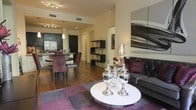 This single-floor residence showcases a thoughtfully designed open-concept layout that effortlessly connects the living, dining, and kitchen areas into one cohesive and inviting space. Dark hardwood flooring runs throughout, lending warmth and a sense of continuity across the entire floor plan.

The living area is anchored by a sleek dark leather sofa adorned with deep plum and burgundy accent pillows, complemented by a round white coffee table styled with elegant silver decorative vessels. A large abstract artwork in charcoal and crimson tones commands the wall, adding a dramatic focal point. A bold area rug in purple and grey grounds the seating arrangement with graphic energy.

The adjacent dining area features a round table with richly upholstered dark red chairs, creating an intimate setting for gatherings. A display shelving unit nearby provides decorative storage with sculptural accessories, adding depth and visual interest.

The kitchen is appointed with dark cabinetry, stainless steel appliances including a full-size refrigerator, a breakfast bar with seating, and pendant lighting overhead. Recessed track lighting throughout the space provides consistent, warm illumination, enhancing the overall sophisticated ambiance.

The cohesive color palette of deep plums, charcoals, and metallics throughout creates a refined and contemporary atmosphere that is both stylish and functional.