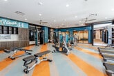 Fitness 44, located at 4401 Wornall Rd in Kansas City, Missouri, is a thoughtfully designed fitness center that combines functionality with modern aesthetics. The space is defined by its striking striped flooring in bold yellow, light blue, and neutral tones, creating an energetic and visually dynamic environment throughout.

The facility is organized across distinct zones, seamlessly connected by black-framed glass partitions and sliding doors that allow natural sightlines while defining separate workout areas. The main cardio zone features a row of Matrix-brand treadmills, stationary bikes, and elliptical machines, all positioned beneath wall-mounted flat-screen televisions displaying vibrant nature scenes. Ceiling fans provide additional air circulation throughout the space.

The strength training area offers a comprehensive selection of equipment, including cable machines, a functional trainer, free weights, benches, and selectorized machines. Large mirrors line the walls, enhancing the sense of openness and allowing users to monitor form during workouts. A dedicated studio space with mirrored walls is accessible via glass doors, suitable for stretching or group exercise.

Near the entrance, a branded reception area with built-in wood cabinetry bears the "Fitness 44" signage, offering a welcoming focal point. Recessed lighting and ceiling fans ensure the space remains well-lit and comfortable, contributing to an inviting, professional fitness environment.
