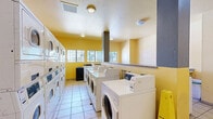 This community laundry facility at 4407 Oakhollow Dr in Sacramento, California, is a well-equipped, single-floor space designed to serve multiple users efficiently. The room is finished with light ceramic tile flooring throughout, contributing to a clean and easy-to-maintain environment. Bright yellow accent walls give the space a lively, welcoming atmosphere, complemented by flush-mount ceiling lights that provide even illumination.

The facility houses a generous number of front-loading washers and stackable washer-dryer units arranged along the walls, offering substantial laundry capacity. A row of top-load machines is also present, providing users with multiple options. Coin-operated or card-based payment terminals are mounted on the machines and walls, streamlining the payment process. A centrally located structural column with a gray exhaust duct serves as a functional architectural element.

Granite-topped folding counters run along one side of the room, offering convenient space for sorting and folding laundry. A series of windows along one wall allows natural light to filter in, enhancing the open feel of the space. Posted instructional signage ensures users can easily navigate machine operation. The overall layout is practical and thoughtfully organized, making this laundry facility a highly functional shared amenity.