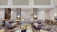 Located at 6805 Walnut Branch Lane in Charlotte, NC, this sophisticated single-floor clubhouse serves as a premier amenity space designed for comfort and social gathering. The interior is defined by soaring double-height ceilings that lend an airy, grand atmosphere throughout.

The lounge area centers around plush, neutral-toned sofas and accent chairs arranged around a glass coffee table, flanked by striking textured column accents in rich, weathered tones. Patterned area rugs and carefully curated décor add warmth and visual interest to the space.

A full-length wall of floor-to-ceiling windows and glass doors floods the interior with abundant natural light while offering direct views of the outdoor pool courtyard. Sheer white drapery softens the light and adds an elegant, airy quality to the room.

The bar and lounge area features an expansive granite-topped island with bar seating, pendant globe lighting, and sleek cabinetry accented by decorative mosaic tile columns. A wall-mounted television and built-in beverage storage make this area ideal for entertaining.

A secondary kitchen lounge includes modern dark cabinetry, stainless steel appliances, gold pendant lighting, and additional bar seating — complemented by curated artwork celebrating the Charlotte skyline. The herringbone-patterned tile flooring ties each area together with a refined, contemporary aesthetic.