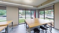 Located at 2323 NE Jackson St in Minneapolis, this single-floor commercial space is thoughtfully designed to support a range of collaborative and functional activities. The layout is divided into several distinct zones, each serving a specific purpose while contributing to a cohesive, modern aesthetic throughout.

The main work area is anchored by a substantial butcher block worktable surrounded by industrial-style stools, creating a versatile surface suited for hands-on tasks or group collaboration. Large floor-to-ceiling windows with roll-down shades line the exterior wall, flooding the space with natural light and offering views of the surrounding neighborhood streetscape. Polished concrete flooring runs throughout, lending a clean, durable foundation that ties the space together.

Adjacent to the work area is a dedicated storage zone featuring rows of matte black individual lockers, providing organized, secure storage for multiple users. Beyond the lockers, a utility kitchen area is visible, equipped with stainless steel surfaces suited for food preparation or equipment use.

A separate lounge area, enclosed by sleek glass walls and a black-framed glass door, offers a more relaxed setting. Inside, bold, colorful murals, a circular pendant light fixture, and comfortable seating create a welcoming, creative atmosphere distinct from the functional work zones.