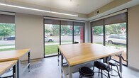 Located at 2323 NE Jackson St in Minneapolis, this single-floor commercial space is thoughtfully designed to support a range of collaborative and functional activities. The layout is divided into several distinct zones, each serving a specific purpose while contributing to a cohesive, modern aesthetic throughout.

The main work area is anchored by a substantial butcher block worktable surrounded by industrial-style stools, creating a versatile surface suited for hands-on tasks or group collaboration. Large floor-to-ceiling windows with roll-down shades line the exterior wall, flooding the space with natural light and offering views of the surrounding neighborhood streetscape. Polished concrete flooring runs throughout, lending a clean, durable foundation that ties the space together.

Adjacent to the work area is a dedicated storage zone featuring rows of matte black individual lockers, providing organized, secure storage for multiple users. Beyond the lockers, a utility kitchen area is visible, equipped with stainless steel surfaces suited for food preparation or equipment use.

A separate lounge area, enclosed by sleek glass walls and a black-framed glass door, offers a more relaxed setting. Inside, bold, colorful murals, a circular pendant light fixture, and comfortable seating create a welcoming, creative atmosphere distinct from the functional work zones.
