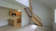 This two-story space is characterized by its open, airy design and practical layout. The main level features light-colored carpeting in the living area, which flows naturally toward the kitchen. The kitchen is equipped with warm honey-toned wood cabinetry, a built-in microwave, an electric range, and light-colored countertops, creating a functional and cohesive cooking space. The cabinetry provides ample storage, and the layout allows for efficient use of the space.

A standout architectural feature is the open-tread wooden staircase, constructed with natural wood and a matching railing system. Its open design allows light to pass through, contributing to the bright, unencumbered feel of the main level. The staircase connects the two floors while adding a visual warmth with its natural wood tones against the neutral white walls.

The overall color palette throughout the space is understated and neutral, with white walls and light flooring creating a clean backdrop. Natural light filters into the living area, enhancing the open atmosphere. The two-floor layout provides a clear separation between living zones, offering both functionality and a sense of spaciousness.