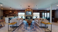 This single-floor community clubhouse or leasing space blends rustic and contemporary design elements to create a welcoming, well-appointed environment. The main lounge area serves as the centerpiece of the space, featuring a striking floor-to-ceiling stacked stone fireplace flanked by a wall-mounted television. Reclaimed wood planks line the accent walls, adding warmth and texture throughout. Two generously sized neutral sofas face each other across a bold, patterned area rug in deep blues and terracotta tones, anchored by a solid wood coffee table. An industrial-style chandelier with exposed Edison bulbs provides ambient overhead lighting, complemented by recessed ceiling lights. Large windows on multiple sides flood the space with natural light and offer direct views of an outdoor pool area, creating a seamless indoor-outdoor connection. Light wood-look flooring runs throughout, tying the design together cohesively. Adjacent to the main lounge is a casual dining nook with a round high-top table and industrial bar stools, a wall-mounted TV, and a dedicated service counter with white cabinetry, quartz countertops, and another reclaimed wood accent wall — ideal for a coffee or refreshment station. The layout flows openly between zones, and glass-paned entry doors provide additional natural light and curb appeal.