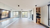 This contemporary fitness studio is thoughtfully designed to support a wide range of movement and wellness activities across a single, well-appointed floor. The space is divided into two primary zones: a bright, open barre and yoga studio, and a fully equipped gym area.

The barre and yoga studio is defined by its clean, minimalist aesthetic. Light hardwood-style flooring runs throughout, complemented by crisp white ceilings with sleek black cylindrical flush-mount fixtures. A wall of floor-to-ceiling mirrors with a mounted barre runs the length of one side, while large black-framed windows on multiple walls flood the space with natural light and offer views of mature trees and the surrounding streetscape. Exposed concrete accent columns add an industrial contrast to the otherwise airy interior. A built-in light wood shelving unit provides organized storage for yoga mats and fitness accessories.

Adjacent to the studio, the gym area is equipped with a multi-functional strength training rig, stability balls, and a variety of exercise equipment, all reflected in mirrored walls that enhance the sense of space. Large windows continue into this zone, maintaining the bright, energizing atmosphere throughout.

Together, the two areas create a cohesive and functional fitness environment that balances form and function with a modern, upscale design sensibility.
