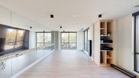 This contemporary fitness studio is thoughtfully designed to support a wide range of movement and wellness activities across a single, well-appointed floor. The space is divided into two primary zones: a bright, open barre and yoga studio, and a fully equipped gym area.

The barre and yoga studio is defined by its clean, minimalist aesthetic. Light hardwood-style flooring runs throughout, complemented by crisp white ceilings with sleek black cylindrical flush-mount fixtures. A wall of floor-to-ceiling mirrors with a mounted barre runs the length of one side, while large black-framed windows on multiple walls flood the space with natural light and offer views of mature trees and the surrounding streetscape. Exposed concrete accent columns add an industrial contrast to the otherwise airy interior. A built-in light wood shelving unit provides organized storage for yoga mats and fitness accessories.

Adjacent to the studio, the gym area is equipped with a multi-functional strength training rig, stability balls, and a variety of exercise equipment, all reflected in mirrored walls that enhance the sense of space. Large windows continue into this zone, maintaining the bright, energizing atmosphere throughout.

Together, the two areas create a cohesive and functional fitness environment that balances form and function with a modern, upscale design sensibility.
