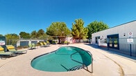 Located at 330 Waymore Dr in El Paso, Texas, this outdoor recreational space centers around a generously sized freeform swimming pool with a smooth, curved design and clearly marked depth indicators. The pool's inviting turquoise water is complemented by a surrounding concrete sun deck that offers ample room for lounging and socializing. Multiple green lounge chairs are arranged around the pool's perimeter, providing comfortable spots for sunbathing beneath El Paso's abundant sunshine. A covered pergola on one side of the deck offers shaded seating, making it a pleasant retreat during warmer months. On the opposite end, a designated picnic area features a table with an umbrella, ideal for casual outdoor dining. The entire pool area is enclosed by chain-link fencing, ensuring both security and privacy. Mature trees line the boundaries of the space, offering natural shade and a touch of greenery against the arid landscape. Adjacent low-rise buildings with a clean, stucco exterior frame the area, housing what appears to be a pool utility or maintenance room with clearly posted safety signage. The overall layout is practical and well-organized, designed to maximize outdoor enjoyment in the sunny West Texas climate.