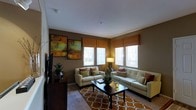 This single-floor space offers a cohesive and thoughtfully designed interior that balances comfort with style. The living room serves as the centerpiece, featuring warm, earthy tones anchored by taupe walls and a plush, patterned area rug in amber and white that adds a bold visual focal point to the room. Two cream-colored sofas are arranged to encourage conversation, accented with rust and green throw pillows that complement the overall warm color palette. A dark wood coffee table sits at the center, adding a grounding contrast to the lighter furnishings. Natural light pours in generously through multiple large windows dressed with woven bamboo-style valances, creating a bright yet cozy ambiance. The windows offer views of the surrounding greenery outside, enhancing the connection to the outdoors. Wall art featuring rich oranges and browns adds a curated, decorative touch above a sleek flat-screen television mounted on the wall. A floor lamp provides additional warm lighting, while potted plants bring a touch of nature indoors. The space exudes a welcoming and harmonious atmosphere, making it an ideal setting for both relaxing and entertaining.