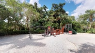 This outdoor recreational space centers around a well-equipped playground set against a dense backdrop of mature tropical trees, including palms and broad-canopied hardwoods that provide natural shade and a verdant setting. The ground throughout the play area is covered in fine, light-colored sand, creating a soft and natural surface suitable for active play.

The main play structure is a multi-component unit finished in red, orange, and green tones, featuring slides, climbing ladders, and decorative sculptural panels that add visual interest. To the left of the central structure stands a set of standalone balance and coordination bars constructed from steel, offering an additional activity zone separate from the main unit. On the right side, a secondary play component extends the overall footprint, incorporating additional climbing and activity elements.

The entire space is framed by dense, mature vegetation that forms a natural green perimeter, lending the area a secluded and park-like atmosphere. The open sky above allows ample natural light to filter through, contributing to a bright and inviting outdoor environment well suited for active recreation.