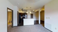 This single-floor space is designed with an open-concept layout that promotes a sense of connectivity and ease of movement throughout. The kitchen serves as a central hub, showcasing dark espresso cabinetry, a tile backsplash, and black appliances including a refrigerator and range. A white breakfast bar peninsula with pendant lighting overhead provides a natural divider between the kitchen and the living area, offering additional counter space and a casual dining option. Track lighting in the kitchen adds a modern, functional touch. The living area features plush carpeting, creating a warm and comfortable contrast to the harder surfaces in the kitchen. White walls and neutral tones throughout keep the space feeling bright and clean. A structural column adds an architectural detail near the transition between spaces. A hallway visible in the background suggests access to additional rooms, providing a logical and well-organized floor plan. The combination of contemporary finishes, thoughtful lighting, and an open layout makes this a practical and inviting single-floor residence.