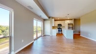This single-floor space is designed with an open-concept layout that seamlessly connects the living area and kitchen, creating a cohesive and functional environment. Warm-toned hardwood-style flooring flows consistently throughout, complementing the neutral gray and taupe wall tones.

The kitchen is a standout feature, outfitted with crisp white shaker-style cabinetry extending to the ceiling for ample storage. Stainless steel appliances — including a refrigerator, electric range, and microwave — are paired with light quartz countertops. A substantial center island provides additional prep space and casual seating potential, while elegant pendant lights hang overhead, adding a refined touch.

The living area is expansive and sun-drenched, benefiting from large sliding glass doors and floor-to-ceiling windows that frame outdoor views of lush greenery. These openings also provide access to a private balcony, extending the livable space outdoors.

The bathroom features a full-size soaking tub and shower combination with clean white surround panels, chrome fixtures, and the same hardwood-style flooring found throughout the unit. The overall ambiance is clean, modern, and move-in ready.