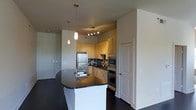 This single-floor space showcases a thoughtfully designed open-concept kitchen that serves as the central feature of the layout. The kitchen is equipped with stainless steel appliances, including a full-size refrigerator and a built-in double oven, complemented by a microwave integrated into the cabinetry. Warm honey-toned wood cabinetry lines the walls, providing ample storage, while a bold mosaic tile backsplash in dark tones adds a striking visual contrast. The kitchen island is a standout feature, finished with a deep charcoal granite countertop and housing a double basin sink with a modern faucet. Overhead, a trio of pendant lights hang at varying heights, casting warm, focused light over the workspace. Track lighting above the kitchen adds additional brightness to the cooking area. Dark hardwood flooring runs throughout, grounding the space with a rich, cohesive tone. The adjacent entryway, visible in the background, features a solid door with a deadbolt, adding a sense of security and separation. Neutral wall tones throughout keep the space feeling clean, open, and versatile.