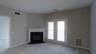 This single-floor space offers a clean, modern layout with a warm and inviting ambiance throughout. The living area serves as the heart of the space, finished with soft, neutral-toned carpeting and walls painted in a calming greige palette that lends a timeless, versatile aesthetic.

A standout feature of the living area is the classic fireplace, framed by a crisp white mantel surround and contrasting black firebox insert, providing both a functional focal point and a refined decorative element. The fireplace adds character and warmth to the space, making it an ideal gathering area.

Natural light flows generously into the room through a set of white-trimmed French doors, which open directly onto a private balcony enclosed by a metal railing. The doors create a seamless visual connection between the interior and the outdoors, enhancing the sense of openness and allowing for excellent ventilation and light.

Practical details include ceiling-mounted HVAC vents and clean white baseboards that run throughout, contributing to the well-maintained and move-in-ready feel of the space. The overall design strikes a balance between comfort and simplicity, making it an adaptable and functional environment suitable for a variety of uses and furnishing styles.