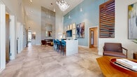 This contemporary single-floor clubhouse is designed to impress, offering a carefully curated blend of functionality and upscale aesthetics throughout its open, flowing layout. The dramatic main lobby commands attention with soaring double-height ceilings, large-format neutral stone tile flooring, and a striking chandelier flanked by pendant lighting. Soft blue-gray walls provide a calming backdrop for bold abstract artwork, while a sleek white reception island with teal velvet bar seating anchors the space. A lounge nook with warm leather armchairs, a wooden slatted accent wall, and a gold side table offers an inviting area for casual conversation. The full kitchen is a standout feature, boasting a large center island with geometric patterned tile detailing, stainless steel appliances, open shelving, and a built-in fireplace set into a floor-to-ceiling tile surround. A dedicated conference room provides a polished setting for meetings, with a live-edge wood table, navy upholstered chairs, a patterned area rug, and ample natural light filtered through roller shades. The fitness center features wall-to-wall floor-to-ceiling windows that flood the space with natural light and outdoor views, alongside modern cardio equipment and wood-look flooring. Every area of this clubhouse reflects thoughtful, contemporary design.