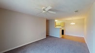 This single-floor unit at Tanyard offers a functional and straightforward layout designed for comfortable everyday living. The living area is generously proportioned, featuring plush wall-to-wall carpeting, neutral-toned walls, and a textured ceiling with a ceiling fan overhead. A sliding glass door fitted with vertical blinds allows natural light to fill the space and provides access to the outdoors. The living area flows openly toward the kitchen, which is separated by a peninsula counter — a practical feature that supports casual dining or food preparation. The kitchen itself is equipped with light wood cabinetry, laminate countertops, hardwood-style flooring, and black appliances including a dishwasher, range, and built-in microwave, alongside a white refrigerator. Ample upper and lower cabinet storage lines the walls. The bedroom features carpeted flooring, a window with blinds that admits natural light, and a textured ceiling with overhead lighting. The bathrooms include a full bath with a tub-shower combo and a second bath featuring a single-sink vanity with light wood cabinetry, a large mirror with bar lighting above, and hardwood-style flooring. The overall ambiance is clean, neutral, and move-in ready.