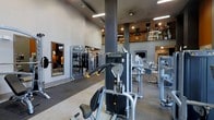This fitness center offers a comprehensive workout environment designed to accommodate a variety of training needs. The space is characterized by its impressive ceiling height, which lends an open, airy feel throughout the main floor. Recessed and overhead lighting ensures the area is well-illuminated, creating a clean and energizing atmosphere.

The main workout floor is outfitted with an array of strength training machines, including cable systems and multi-function equipment arranged to allow comfortable movement between stations. A mirrored wall section along one side enhances the sense of space and allows users to monitor their form during exercise. The flooring transitions between rubberized and hard surfaces, delineating functional training zones.

A mezzanine-level area is visible overhead, featuring additional equipment and what appears to be a dedicated cardio zone, expanding the overall capacity of the facility. Structural columns define the space without impeding the flow of movement. The neutral color palette of grays and whites, combined with warm accent tones on the upper level, contributes to a modern and professional aesthetic throughout.