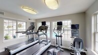 This well-equipped fitness center in Florissant, Missouri offers a comprehensive workout environment designed to support a wide range of exercise routines. The single-floor facility is thoughtfully laid out, dividing the space into distinct cardio and strength training zones.

The cardio area is lined with multiple Life Fitness treadmills and elliptical machines, each equipped with individual monitor screens. Large windows along the walls flood the space with natural light and provide views of the adjacent outdoor pool area, creating an energizing atmosphere for endurance training.

The strength training zone features a Life Fitness multi-functional cable machine with suspension training attachments, a dedicated free weight rack with a selection of dumbbells, and a weight bench, offering a solid foundation for resistance training. Full-length mirrors are mounted along one wall, allowing users to monitor their form during exercises.

Durable rubber flooring covers the entire facility, providing safety and shock absorption throughout. Overhead, large circular flush-mount light fixtures provide consistent, bright illumination. The space also includes a convenience station near the entry door with signage and storage. A glass-panel exit door opens directly to the exterior, ensuring easy access. The facility is monitored by video surveillance for added security.