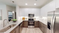 This well-designed single-floor residence showcases a thoughtful layout and contemporary finishes throughout. The kitchen is a standout feature, boasting a two-tone cabinet design with crisp white upper cabinets and warm taupe lower cabinets, complemented by granite countertops, a textured tile backsplash, and stainless steel appliances including a range, microwave, and dishwasher. Under-cabinet lighting adds a refined touch, while light wood-style flooring ties the space together seamlessly. A dedicated dining area sits adjacent to the kitchen, bathed in natural light from a glass door and framed by track lighting overhead — a welcoming spot for everyday meals or entertaining. The bedroom is generously sized, featuring plush carpeting, a ceiling fan, and a window that allows natural light to filter in. The en-suite bathroom is well-appointed with dual granite-topped vanities, a large mirror, and a soaking tub and shower combination finished with neutral tile. Directly accessible from the bathroom is a walk-in closet with built-in shelving, offering ample storage. Throughout the residence, clean white walls, recessed lighting, and consistent flooring create a cohesive and modern aesthetic.