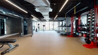 This well-appointed fitness center offers a comprehensive workout environment designed to accommodate a wide range of exercise needs. The space is characterized by an industrial-inspired aesthetic, with an exposed black ceiling revealing visible HVAC ductwork and structural elements, complemented by sleek linear LED lighting that ensures bright, even illumination throughout.

A defining architectural feature is the expansive floor-to-ceiling window wall along one side of the space, flooding the interior with natural light and providing open views of the surrounding urban streetscape. The durable, cushioned rubber flooring in a warm tan tone covers the entire area, providing both comfort and safety for all types of training.

The cardio zone is well-equipped with multiple treadmills and elliptical machines positioned to take advantage of the exterior views. A dedicated strength training area includes a Precor cable machine, a rack of dumbbells in varying weights, and weight benches. A standout functional training station features a bold red rig equipped with TRX suspension trainers, kettlebells, medicine balls, and battle ropes. Wall-mounted mirrors expand the sense of space and aid in form monitoring. A water fountain and additional utility amenities are also present, rounding out this fully functional fitness facility.
