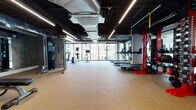 This well-appointed fitness center offers a comprehensive workout environment designed to accommodate a wide range of exercise needs. The space is characterized by an industrial-inspired aesthetic, with an exposed black ceiling revealing visible HVAC ductwork and structural elements, complemented by sleek linear LED lighting that ensures bright, even illumination throughout.

A defining architectural feature is the expansive floor-to-ceiling window wall along one side of the space, flooding the interior with natural light and providing open views of the surrounding urban streetscape. The durable, cushioned rubber flooring in a warm tan tone covers the entire area, providing both comfort and safety for all types of training.

The cardio zone is well-equipped with multiple treadmills and elliptical machines positioned to take advantage of the exterior views. A dedicated strength training area includes a Precor cable machine, a rack of dumbbells in varying weights, and weight benches. A standout functional training station features a bold red rig equipped with TRX suspension trainers, kettlebells, medicine balls, and battle ropes. Wall-mounted mirrors expand the sense of space and aid in form monitoring. A water fountain and additional utility amenities are also present, rounding out this fully functional fitness facility.