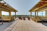 Located at 16 Cinema Dr in Lansing, NY, this rooftop terrace offers an expansive outdoor retreat designed for relaxation and social gathering. The space is defined by its open, tiled terrace deck, bordered by sleek horizontal metal railings that provide unobstructed panoramic views of the rolling hills, lush greenery, and open skies characteristic of the Finger Lakes region.

Two substantial wood-framed pergolas anchor opposite sides of the terrace, providing partial shade while preserving the airy, open-air atmosphere. Beneath each pergola, built-in wooden bench seating and picnic-style tables create comfortable gathering nooks ideal for conversation or al fresco dining. Raised wooden planter boxes line the perimeter, adding greenery and a warm, natural aesthetic to the space.

At the center of the terrace, a cluster of classic Adirondack chairs invites visitors to sit back and take in the sweeping views. The large-format tile flooring ties the entire space together, lending a clean, modern finish to the rooftop. The overall ambiance is bright, welcoming, and open, making this terrace a standout communal amenity suited for enjoying warm-weather days and the scenic natural surroundings of Lansing, New York.