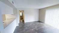 This single-story space offers a clean, straightforward layout designed for comfortable everyday living. The main living area is finished with durable wood-style plank flooring in a rich, dark tone that contrasts neatly with the crisp white baseboards and neutral beige walls, creating a warm yet understated aesthetic throughout. A textured popcorn ceiling adds a classic touch overhead, while the open wall space provides flexibility for a variety of furniture arrangements. A hallway visible from the main room leads to additional areas of the home, with natural light streaming in through a window at the far end, enhancing the sense of depth and openness. The doorway framing and architectural transitions between rooms are clean and simple, maintaining a cohesive look throughout the space. The neutral color palette—spanning soft beige walls, white trim, and dark flooring—creates a versatile backdrop that accommodates a wide range of interior design styles. Overall, this space presents a move-in-ready environment with practical finishes and a well-considered layout suited for a variety of occupants.
