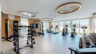 Located at 200 Daytona Blvd in Daytona Beach, FL, this contemporary fitness center is a well-appointed, single-floor facility offering a comprehensive workout environment. The space is divided into two distinct zones designed to accommodate a full range of exercise needs.

The main strength and cardio area features an impressive array of equipment, including treadmills, ellipticals, stationary bikes, a Smith machine, and a full rack of dumbbells. A large flat-screen television is mounted on the wall, and floor-to-ceiling windows line the perimeter, flooding the space with natural light and offering views of the surrounding Daytona Beach landscape, including palm trees and the colorful cityscape beyond.

A secondary functional training area provides an open, flexible floor plan ideal for dynamic workouts. This zone is equipped with medicine balls, kettlebells, and plyometric boxes, and benefits from the same expansive window views and abundant natural light. Large sliding glass doors lead directly outside, enhancing the airy atmosphere.

Throughout the facility, the design aesthetic is sleek and modern, defined by warm wood slat accent walls, full-length mirrors, durable wood-tone flooring, and signature oval-shaped recessed lighting fixtures that lend the space a sophisticated, boutique-gym ambiance.
