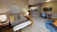 This single-floor residence offers a thoughtfully designed open-concept layout that connects the living, dining, and kitchen areas into one cohesive and functional space. The living area is warm and inviting, featuring plush carpeted flooring, neutral taupe walls, and ample room for comfortable furnishings. A textured area rug anchors the seating arrangement, which is complemented by decorative accent pillows and a sleek marble-top coffee table that adds a refined touch to the space. A large framed artwork on the wall adds a contemporary artistic element. The kitchen is situated on a raised platform and features white cabinetry, white appliances, and a breakfast bar-style peninsula that provides both workspace and a casual dining option. Track lighting overhead ensures the kitchen is well-lit and practical. Adjacent to the living area, a designated dining space offers room for a table and chairs, making it ideal for everyday meals or entertaining. The open flow between all areas creates a sense of spaciousness and versatility, while the consistent warm, neutral color palette throughout lends the space a cohesive and welcoming ambiance. The overall design balances simplicity and comfort, making it a highly livable and functional home.
