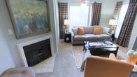 This single-floor living space is thoughtfully designed to balance comfort and style. The main living area serves as the focal point of the home, anchored by a classic fireplace with a tiled hearth and a bold, large-scale artwork displayed above the mantle, adding a striking visual element to the room. The space is furnished with a plush, neutral-toned sofa accented by warm orange and teal throw pillows, complemented by a coordinating armchair that reinforces the cohesive color palette. A glass-topped coffee table sits atop a geometric-patterned area rug, adding a layer of texture and visual interest to the carpeted floor. Natural light filters through large windows dressed in eye-catching patterned curtains featuring bold orange and teal tones, which tie the room's color scheme together seamlessly. A ceiling fan with a light fixture overhead provides both comfort and illumination. A cylindrical black side table and a stylish table lamp complete the seating arrangement, offering both functionality and decorative appeal. The overall ambiance is warm and curated, with a strong sense of personal style evident in every detail of the space.