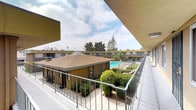 This residential complex is organized around a central courtyard that serves as the heart of the community. The buildings are finished in a distinctive gold and brown color scheme, giving the property a cohesive and recognizable character. A generously sized rectangular swimming pool anchors the courtyard, enclosed by a metal security fence and surrounded by a concrete pool deck furnished with lounge chairs — ideal for outdoor relaxation. The pool area is framed by neatly landscaped greenery, including shrubs, flowering plants, and mature trees that provide shade and a sense of privacy. Open-air walkways run along the upper level of the buildings, offering residents direct balcony-style access to their units while providing elevated views of the courtyard and surrounding neighborhood. Exterior staircases connect the upper corridor to the ground level, facilitating convenient movement throughout the property. A central structure within the courtyard, featuring a pitched roof, appears to serve as a shared facility or leasing office. The overall layout promotes a communal atmosphere, with the well-maintained landscaping and shared amenities contributing to an inviting outdoor environment.