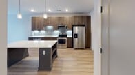 Located in Durham, NC, this modern single-floor residence showcases a thoughtfully designed open-concept layout with clean lines and contemporary finishes throughout. The kitchen serves as a central hub, featuring rich wood-toned cabinetry, a light quartz countertop island with built-in shelving, a glass tile backsplash, and a full suite of stainless steel appliances including a refrigerator with ice and water dispenser, range, microwave, and dishwasher. Pendant lighting above the island adds warmth and visual interest. The kitchen flows seamlessly into the living and dining areas, both finished with light-toned hardwood-style flooring that extends throughout the main living spaces. Large windows dressed with horizontal blinds fill these areas with natural light, enhancing the open and airy feel. The bedroom offers plush wall-to-wall carpeting, a ceiling fan, and a generously sized window overlooking the outdoors. The bathroom is elegantly finished with light wood flooring, warm wood cabinetry paired with matte black hardware, a white quartz vanity, and a standout glass-panel shower enclosure framed in bold black metal. The overall aesthetic is modern and cohesive, combining neutral tones with carefully selected accent finishes for a polished, move-in ready appeal.