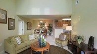 This two-story residence offers an inviting and well-connected layout that blends comfort with practicality. The main living area is anchored by a spacious sitting room with a plush sofa, armchair, and a round wooden coffee table, all set atop a rich, dark area rug. The room benefits from soaring double-height ceilings that create an airy, open atmosphere, enhancing the sense of volume and space throughout.

Flowing seamlessly from the living room is a dedicated dining area, furnished with a wooden table and chairs, accented by a pendant light above — ideal for everyday meals and gatherings. The kitchen sits adjacent, separated by a half-wall counter that maintains visual connectivity. It features warm wood cabinetry, white appliances including a range, and overhead lighting, offering a functional and efficient cooking space.

Throughout the main level, neutral-toned walls in soft green hues and light-colored carpeting contribute to a calm and cohesive aesthetic. Decorative touches such as wall art, potted plants, and layered accessories add warmth and personality to the space. The open sightlines across the living, dining, and kitchen areas make the home feel welcoming and easy to navigate, while the second floor adds additional living or sleeping accommodations above.