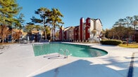 Located at 6147 Winged Elm Ct in Charlotte, NC, this outdoor amenity area serves as a central gathering hub for the residential community. The centerpiece is a generously sized resort-style swimming pool with a dedicated wading area, set against a backdrop of tall mature pine trees and the community's distinctive red and beige multi-story residential buildings. The pool deck is finished in smooth concrete and offers ample space for lounging and recreation.

Surrounding the pool, the sundeck is outfitted with multiple lounge chairs and seating sets shaded by a teal patio umbrella, creating a comfortable outdoor retreat. The area enjoys abundant natural sunlight, making it an inviting space throughout the day.

Adjacent to the pool is a single-story clubhouse building clad in light beige lap siding, featuring large multi-pane windows that provide clear sightlines to the pool area. The clubhouse entrance is marked by double glass doors and a Bridge Property Management welcome mat, indicating professionally managed facilities. Pool rules are clearly posted at the building exterior. The overall ambiance is clean, open, and well-maintained, offering residents a welcoming outdoor environment designed for relaxation and community enjoyment.