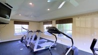 This single-floor fitness space is designed to support a focused and comfortable workout environment. The room is outfitted with multiple cardio machines, including treadmills and an elliptical trainer, all positioned on durable rubber flooring suited for high-impact exercise. Two ceiling fans are mounted overhead, providing consistent airflow throughout the space and contributing to a comfortable atmosphere during workouts. Natural light pours in generously through several large, multi-pane windows dressed with striped Roman shades, offering views of the surrounding greenery outside. The warm, neutral-toned walls complement the clean, functional aesthetic of the room. Recessed lighting supplements the natural light, ensuring the space remains well-lit at any time of day. The layout is thoughtfully arranged to allow ease of movement between equipment. Overall, this fitness room combines practical design with a bright, airy ambiance, making it a welcoming and functional space for regular exercise.