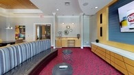 The Media Center is a thoughtfully designed, single-floor space that seamlessly blends entertainment, relaxation, and collaborative functionality. The interior is immediately distinguished by its striking crimson and charcoal geometric-patterned carpeting, which anchors the entire space with a bold visual energy.

The main lounge area features a curved, striped sofa complemented by sleek black leather swivel chairs and low-profile round side tables, creating an inviting setting for casual gatherings. A dedicated media wall, framed in warm blonde wood with built-in speakers and display units, serves as the entertainment focal point and is paired with ample credenza storage below.

Natural light filters in through large windows dressed with floor-length drape panels, offering views of the surrounding greenery. A recessed wood-paneled ceiling detail adds warmth and architectural interest overhead, while recessed lighting and pendant fixtures provide layered illumination throughout.

A separate alcove accommodates a round meeting table with ergonomic chairs beneath a statement pendant light and vibrant abstract artwork, making it ideal for small group work or discussions. A fireplace with a built-in display shelf and an adjacent settee rounds out the space, contributing to its versatile, comfortable ambiance. Crown molding and structural columns add classic architectural refinement throughout.